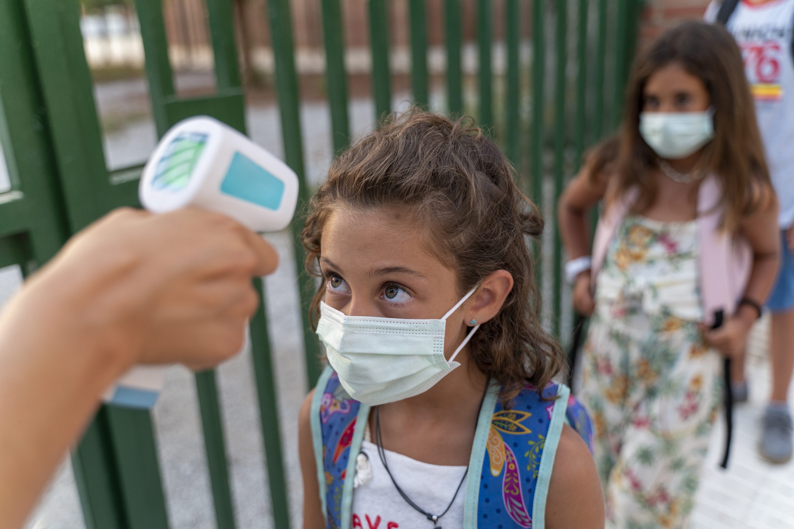 Teacher woman checking temperature of students using a laser thermometer at the school door; kids in line wearing masks.