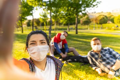 College age woman taking selfie with big smile wearing a mask. Two college-aged men are sitting on the grass behind her