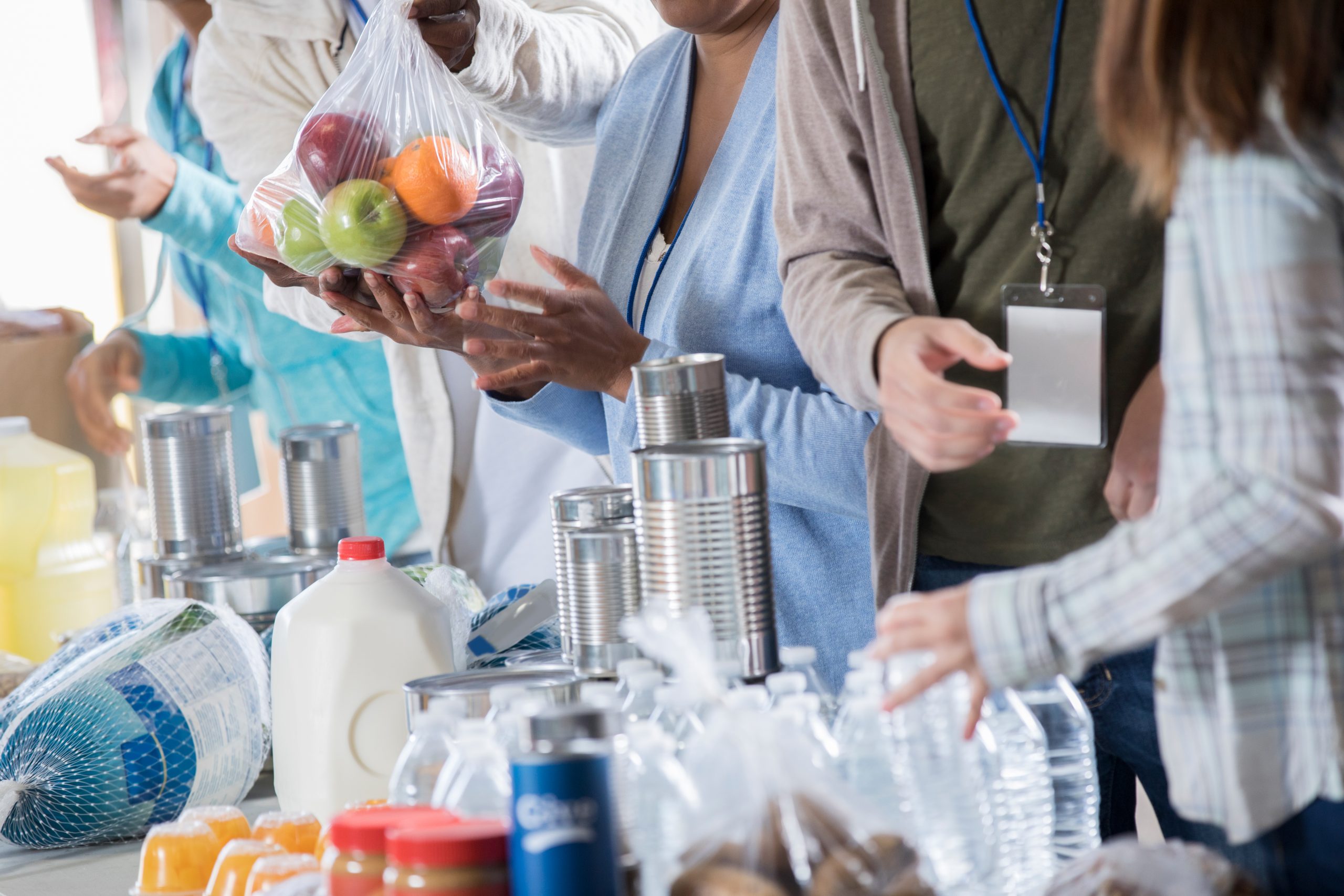 Group of unrecognizable people volunteer at a food bank. They are receiving donated canned food, fruit, milk and other food staples