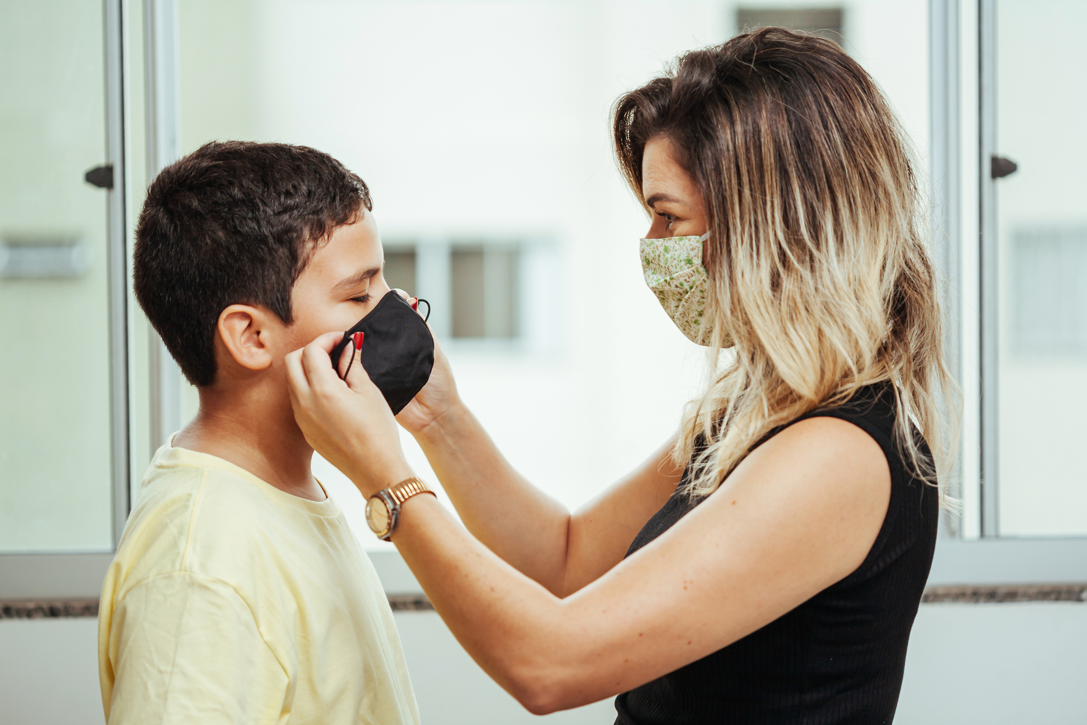 Mother wearing white and green cloth face mask faces son while putting on black coth fask mask on this face.