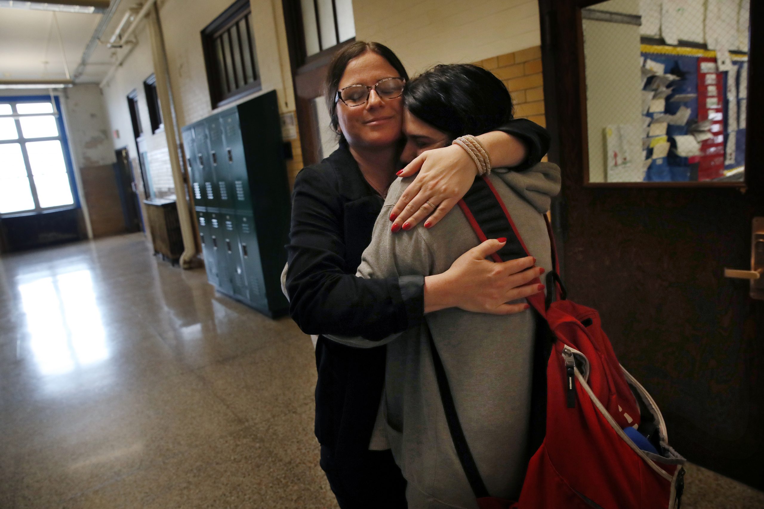 Female Counselor hugs female student with red backpack in high school hallway