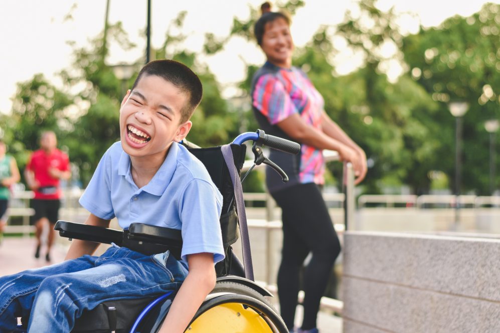 child on wheelchair wearing light blue polo shirt and jeans is laughing in outdoor park