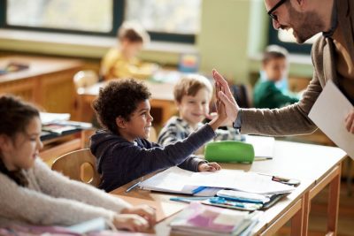 a male teacher gives a young boy a high five in a classroom. They are both smiling while other students look on.