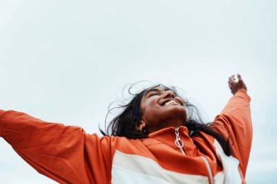 A person is seen from behind standing outdoors, wearing a bright orange and white windbreaker-style jacket with both arms raised high against a clear blue sky. The lighting and posture convey a sense of triumph, freedom, or celebration. The image evokes a feeling of optimism and victory.
