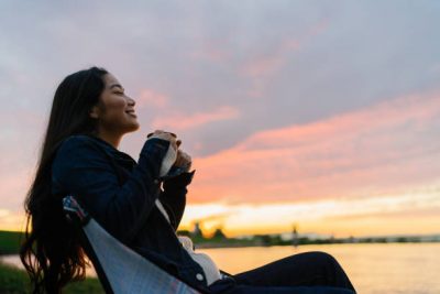 A young woman is enjoying sitting near a lake and drinking a hot drink in nature during sunset.
