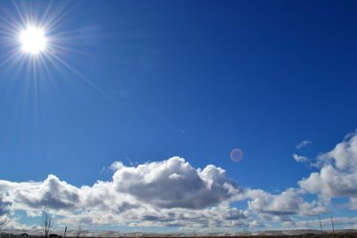 A bright sun shines in the top left corner of a clear blue sky, casting rays of light. Fluffy white clouds are scattered across the horizon.