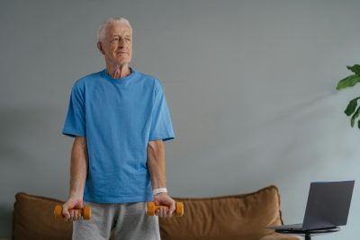 An older adult standing in a living room holding two small orange dumbbells. They are dressed in a blue T-shirt and gray sweatpants. Behind them is a brown couch, and to the right, a small table holds an open laptop. A green plant is partially visible on the far right side of the image.