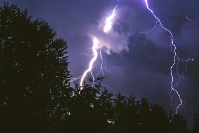 A nighttime scene with a dark sky lit up by several bright lightning bolts. Tree silhouettes are visible in the foreground, and the lightning appears to strike through and around them. The sky features a mix of dark clouds and glowing areas from the lightning.