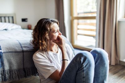 A woman sits on the floor at the foot of a bed with a worried look on her face. She rests her face on her hand.