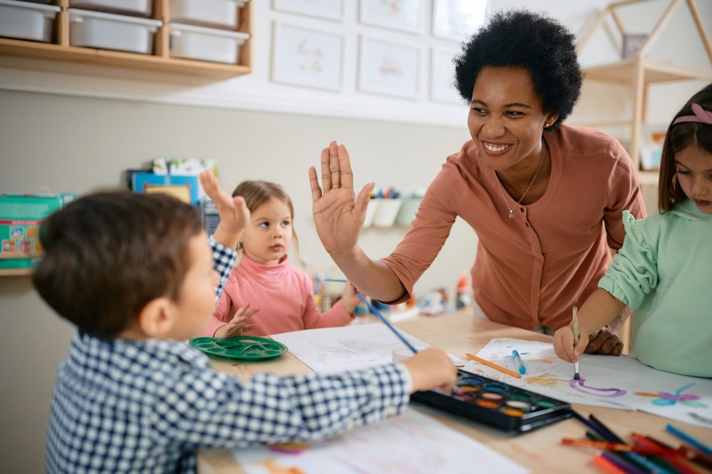 A teacher smiles and gives a young boy high five in the classroom while two classmates look on